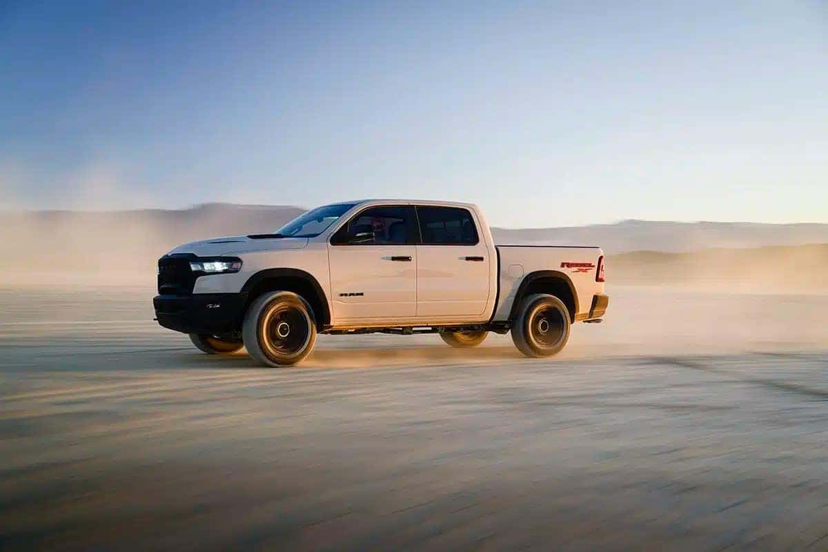 White RAM 1500 Rebel X driving fast across a desert plain, kicking up dust with mountains in the background and golden hour lighting.
