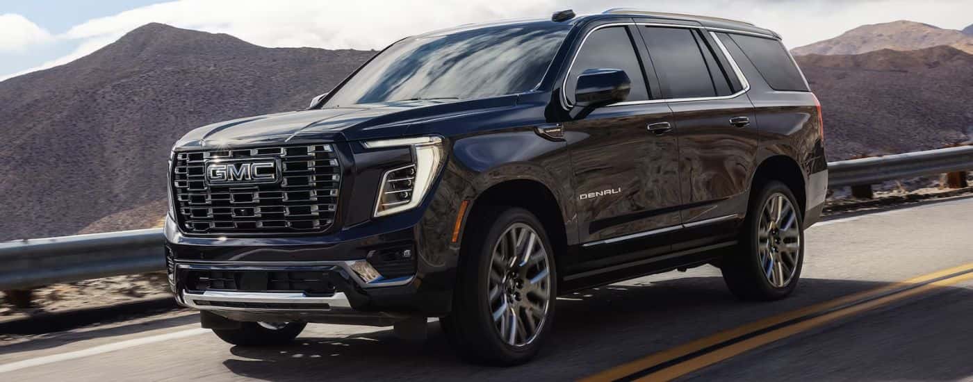 A black 2026 GMC Yukon Denali driving down a road with mountains in the background.