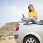 Woman in Yellow Blouse and Blue Jeans Taking Selfie While Sitting on Car