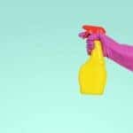 A teal background with a hand in a pink latex glove and a spray bottle popping out from the right side.