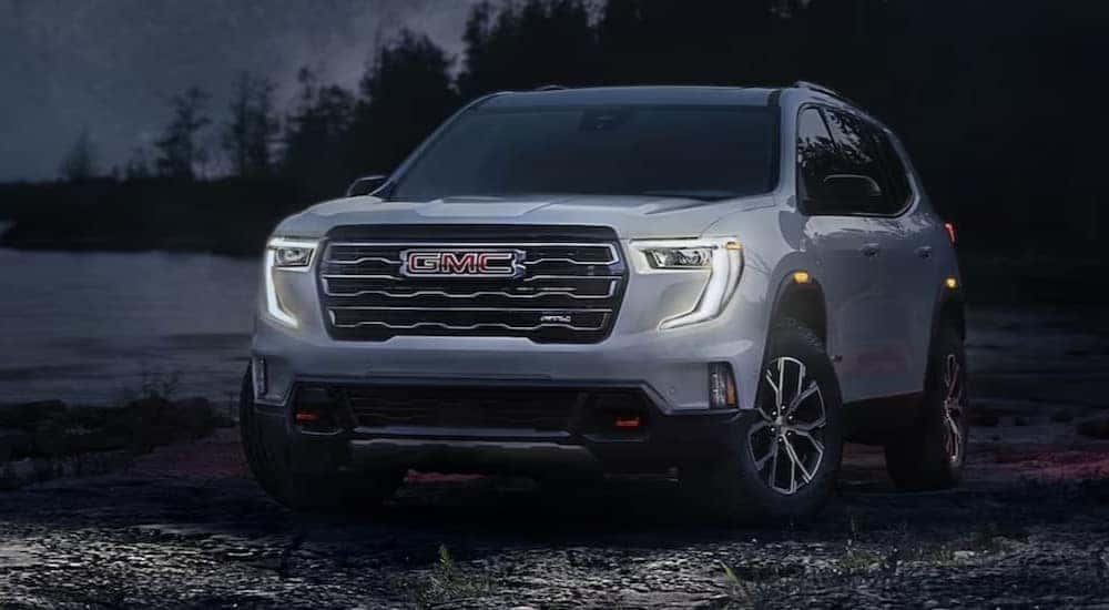 A silver 2024 GMC Acadia AT4 is shown parked with the headlights on.