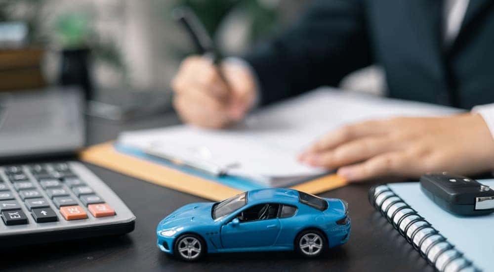 Close-up of a blue toy car sitting on a desk while a person writes on a clipboard.