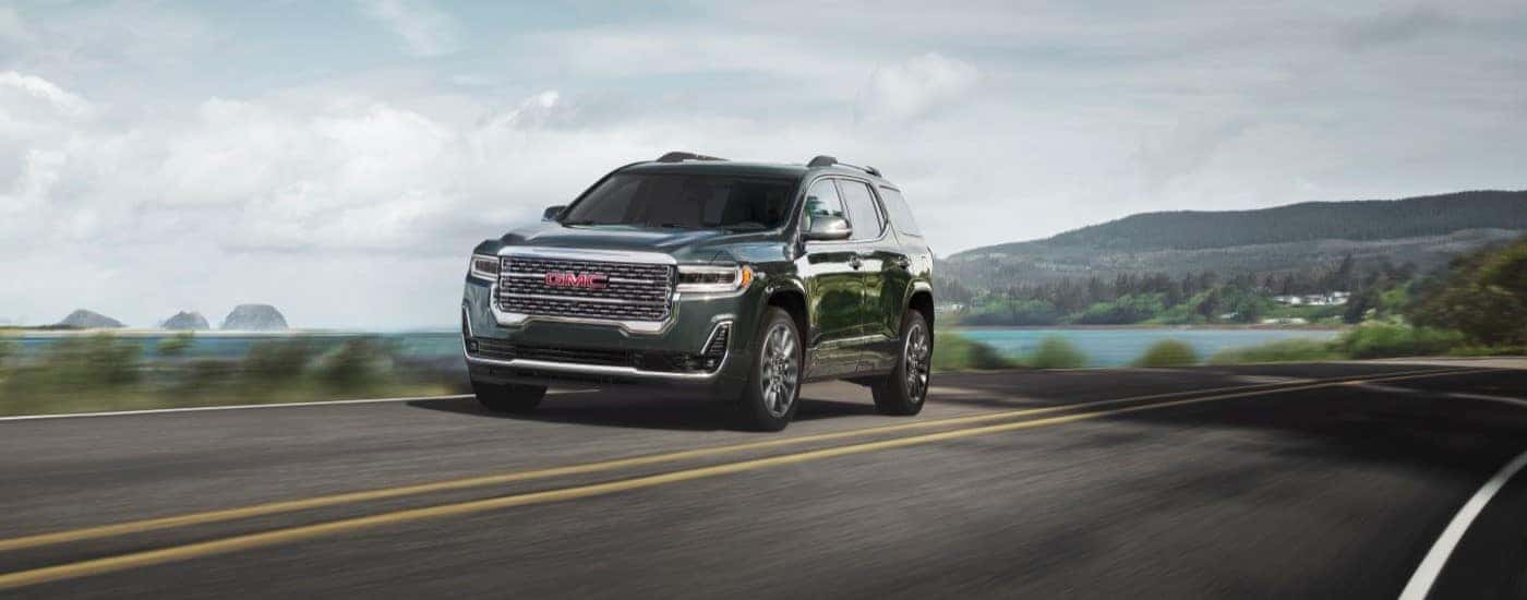 A black 2023 GMC Acadia Denali is shown driving past a laker and mountains.