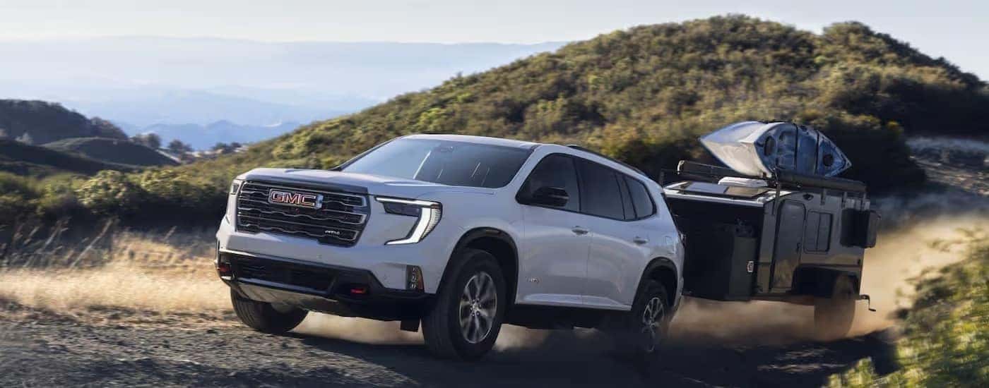A white 2025 GMC Acadia AT4 is towing a small camper on a dirt road.