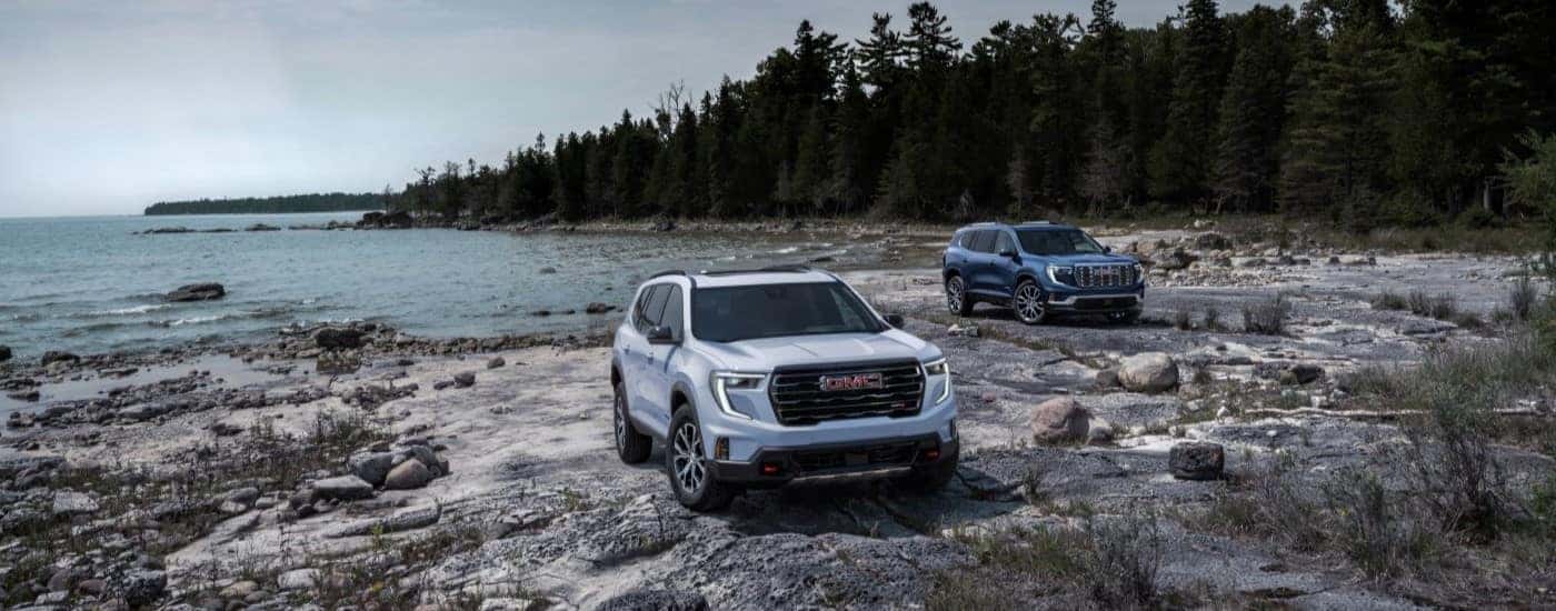 A white 2024 GMC Acadia parked next to a blue 2024 GMC Acadia parked by water