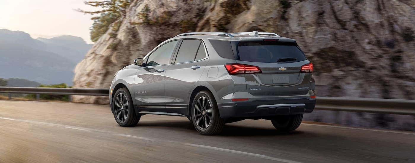 A grey 2023 Chevy Equinox Premier is shown from a rear angle driving past a rock face.