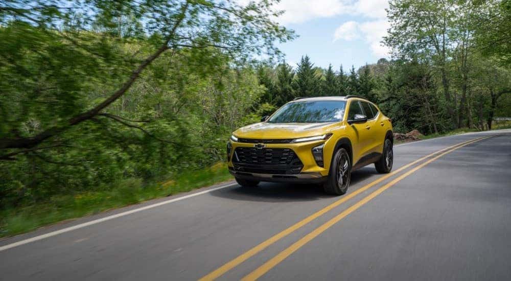A yellow 2024 Chevy Trax Activ is shown driving on a tree-lined road after leaving a Chevy dealer near Duluth.