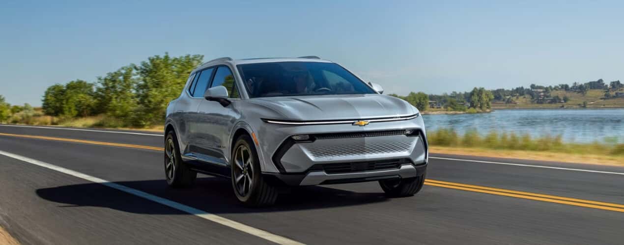 A silver 2025 Chevy Equinox EV is shown driving past a lake.