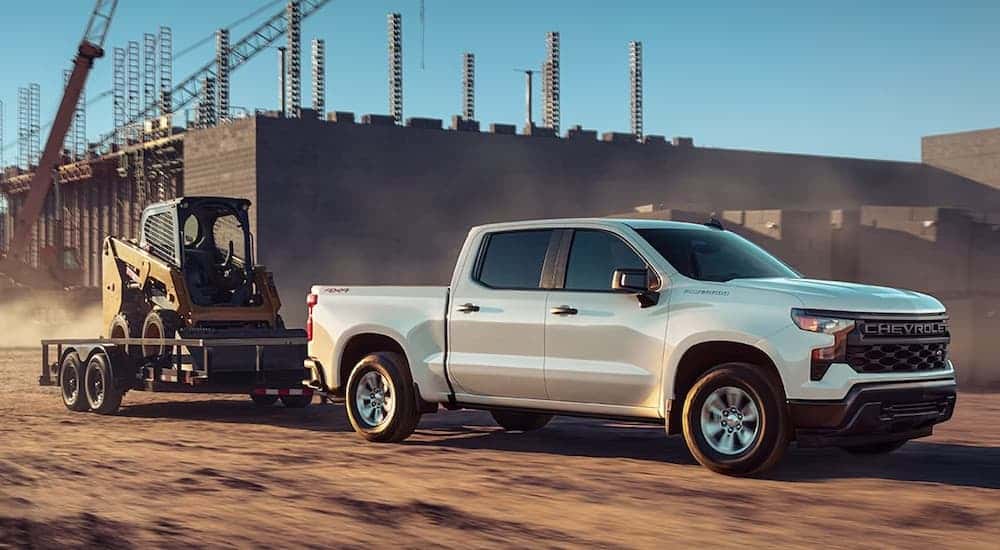 A white 2025 Chevy Silverado 1500 from the side while towing a skid-steer.
