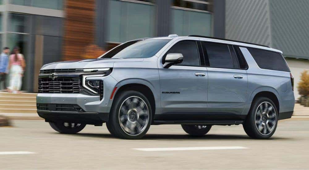 Silver 2026 Chevy Suburban LS driving past a modern apartment building.
