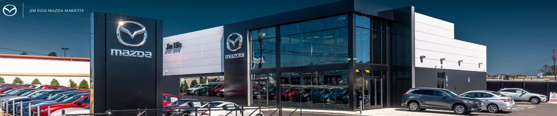 Exterior daytime view of a Mazda dealership showroom with black and white facade architecture and cars parked outside.