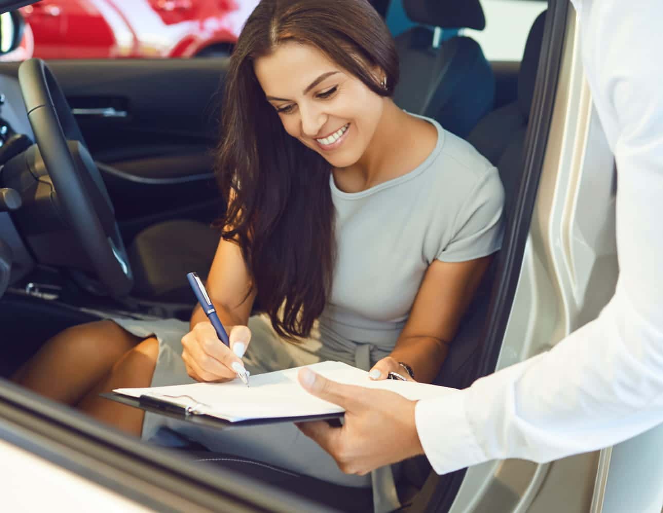 Woman sitting inside a vehicle signing paperwork held by a salesperson.