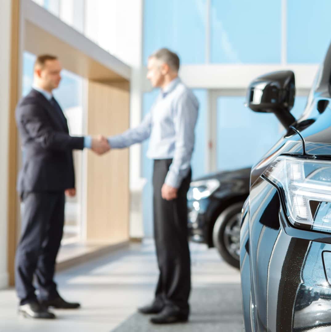 Two men shaking hands inside a car dealership with a vehicle in the foreground.