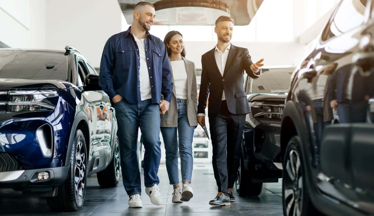 A couple walking through a car dealership showroom with a sales representative who gestures toward vehicles on display.