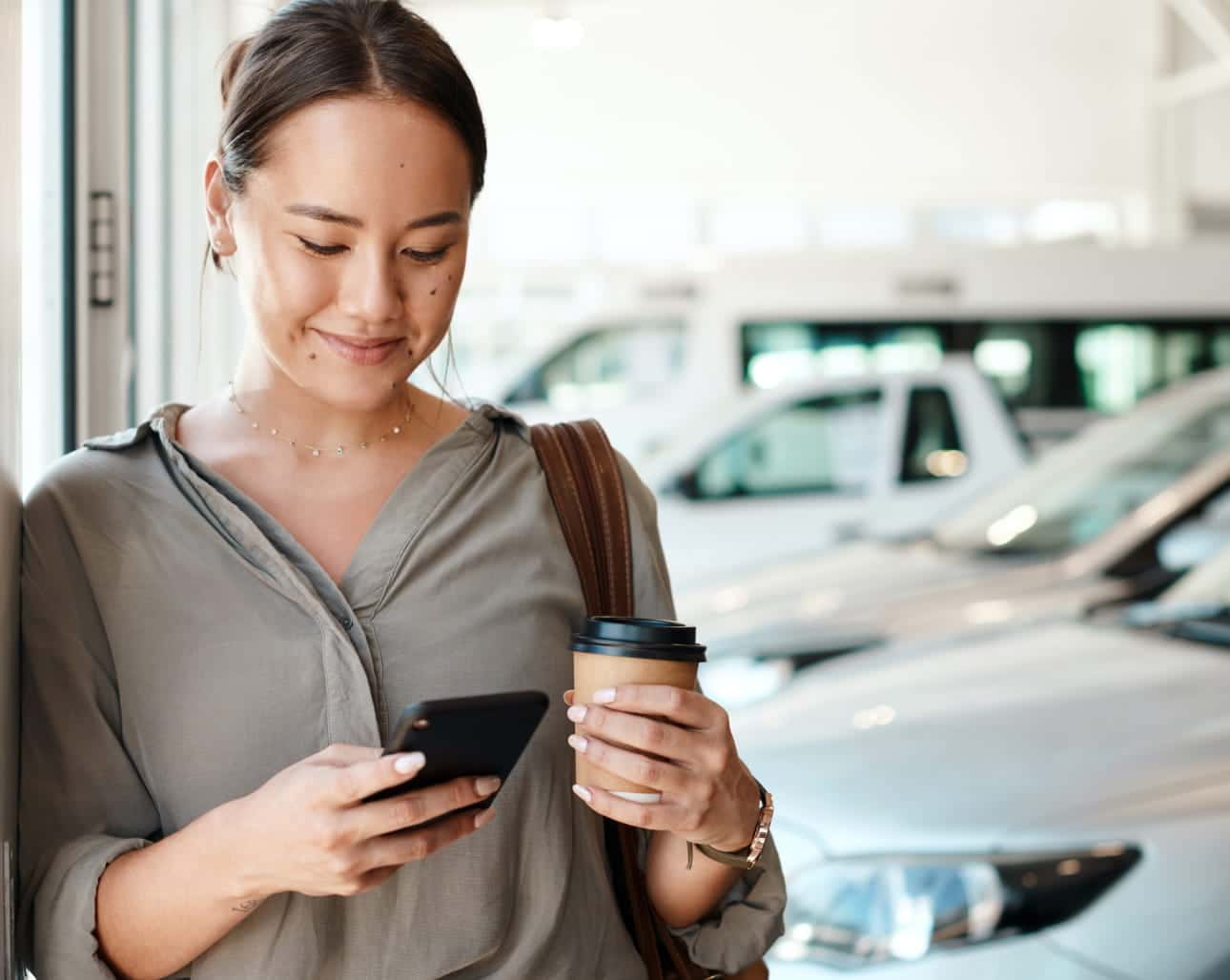Woman holding a coffee cup while using her smartphone inside a bright car dealership showroom with vehicles in the background.