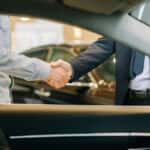 A car salesperson and customer shaking hands through a vehicle's window