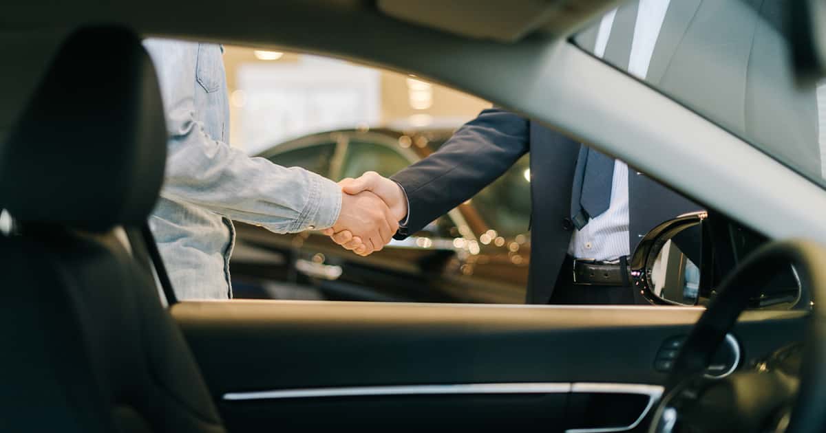 A car salesperson and customer shaking hands through a vehicle's window