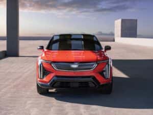 2026 Cadillac OPTIQ, Sport 2 (6MR26), exterior detail, red, Monarch Orange (GAG), 21 Inch wheels (RVU), front view, head on, parked, parking structure, rooftop, city, skyline, sunset, concrete pavement, building, cloudy;