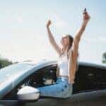 Happy teen girl sitting on the windowsill of a parked car, raising her arms in celebration and holding car keys, excited about getting her first car on a sunny day.