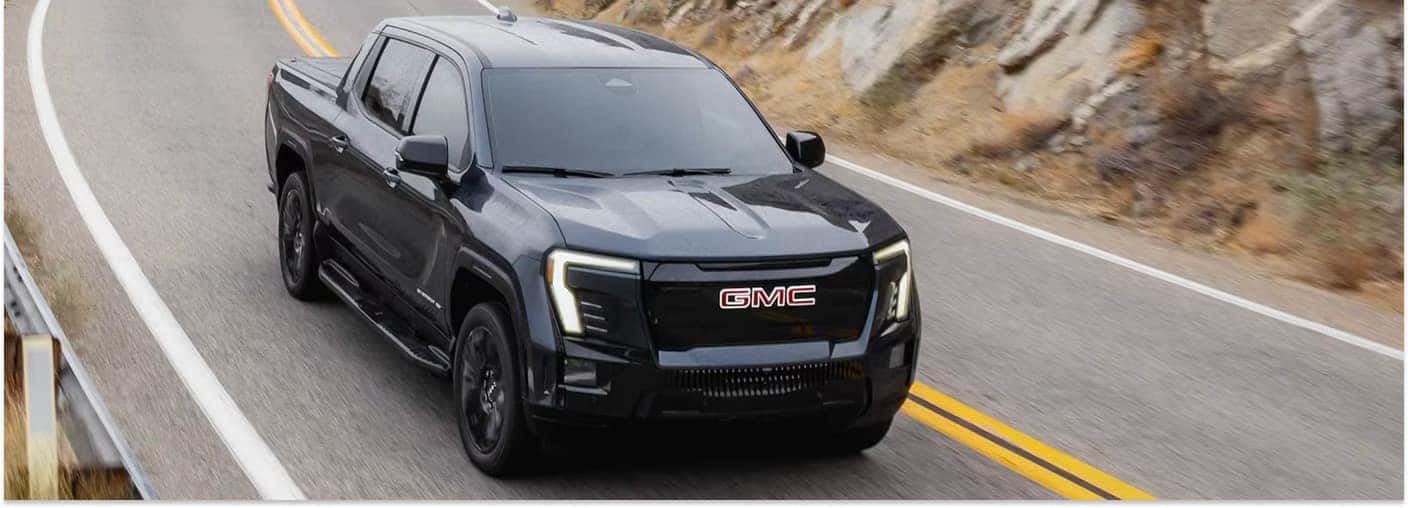 A dark gray 2026 GMC Sierra EV pickup truck navigates a curved asphalt road, set against a backdrop of a rocky hillside.