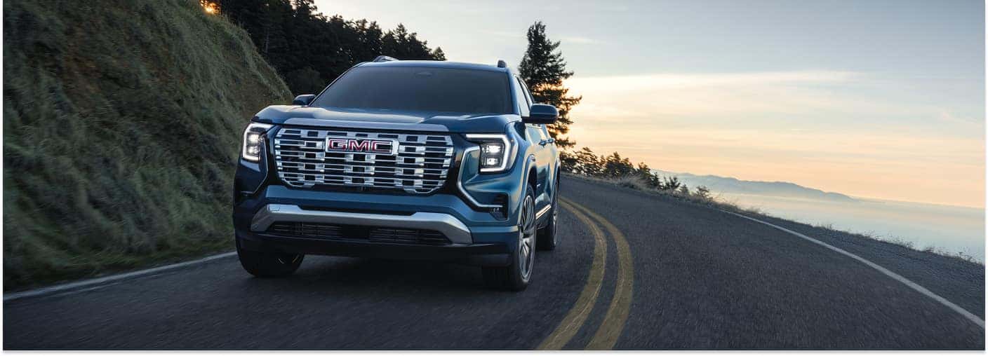 A blue 2026 GMC Terrain SUV drives along a winding coastal road, set against a scenic backdrop of a misty ocean overlook at sunset.