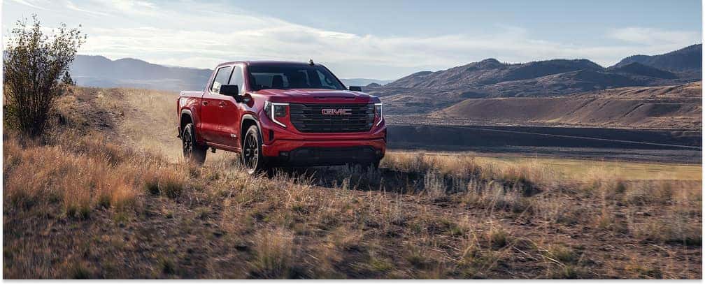 Red 2026 GMC Sierra 1500 pickup truck driving on an off-road dirt trail with a mountain backdrop.