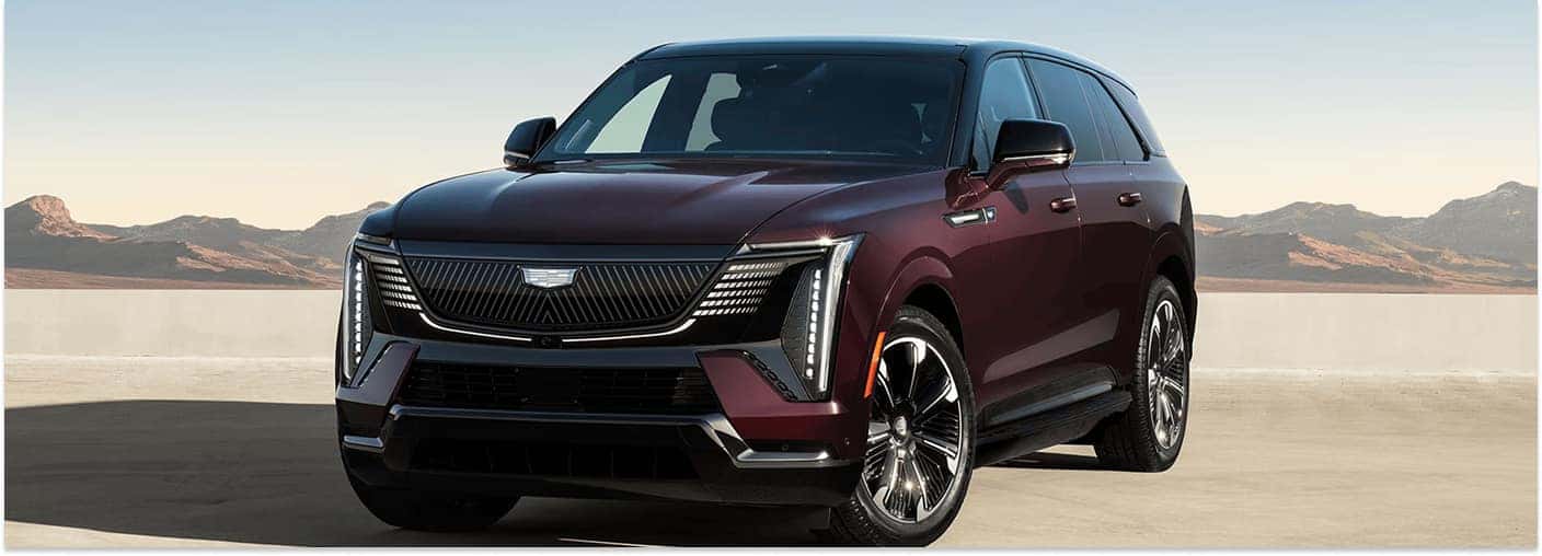 A front three-quarter view of a deep burgundy 2026 Cadillac Escalade IQ parked on a flat concrete surface with a desert mountain landscape in the background.