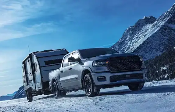 Silver 2027 Ram 1500 towing a camper trailer through snowy mountains