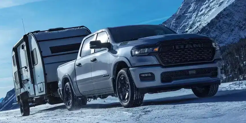 Silver 2027 Ram 1500 towing a camper trailer through snowy mountains