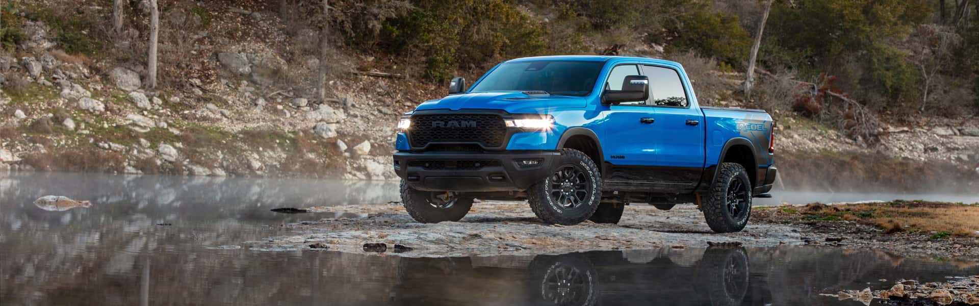 2025 1500 Rebel in two-toned Hydro Blue and Diamond Black with daytime running lights on, parked on a foggy riverbank surrounded by water, trees and cloudy gray skies.