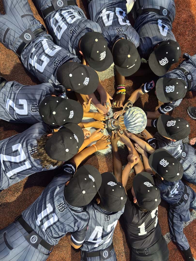 kids in baseball attire laying down around a trophy with their hands touching the trophy