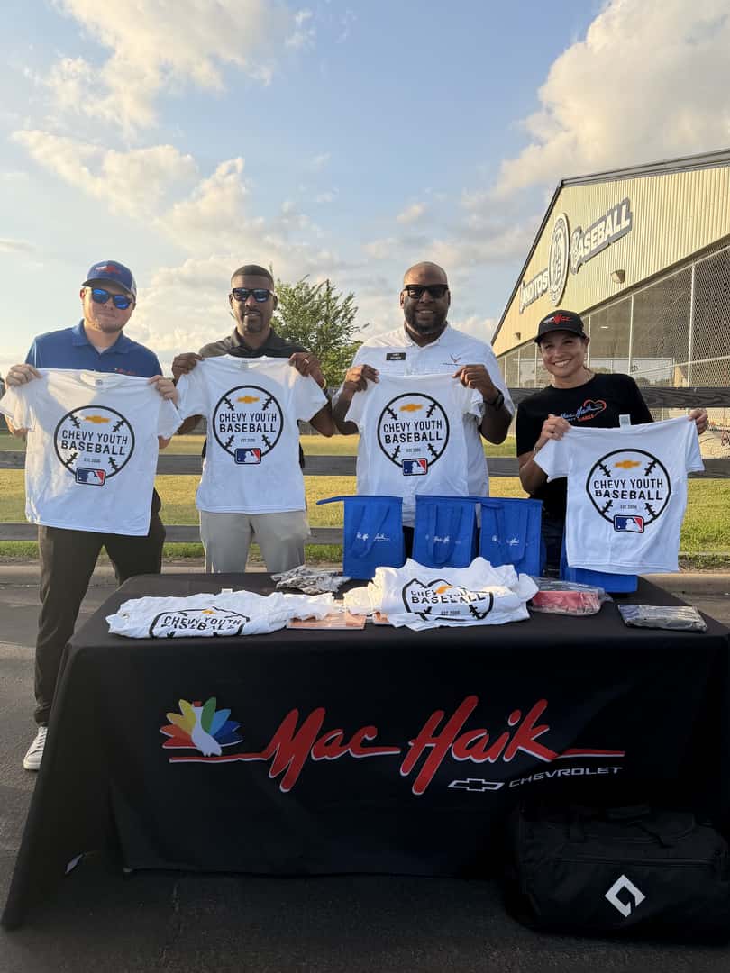 Four people holding up shirts for the Mac Haik Chevy Youth Baseball charity