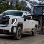 2026 GMC Sierra HD towing a massive tracked all-terrain vehicle on a flatbed trailer down a dusty mountain dirt road, with snowy peaks in the background.