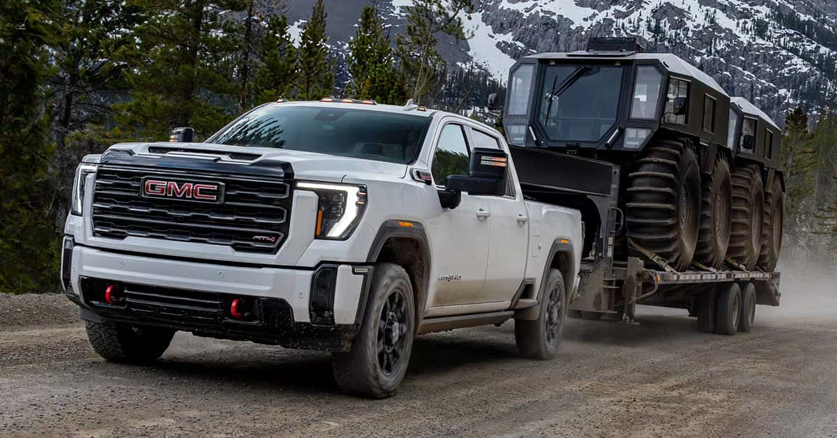 2026 GMC Sierra HD towing a massive tracked all-terrain vehicle on a flatbed trailer down a dusty mountain dirt road, with snowy peaks in the background.