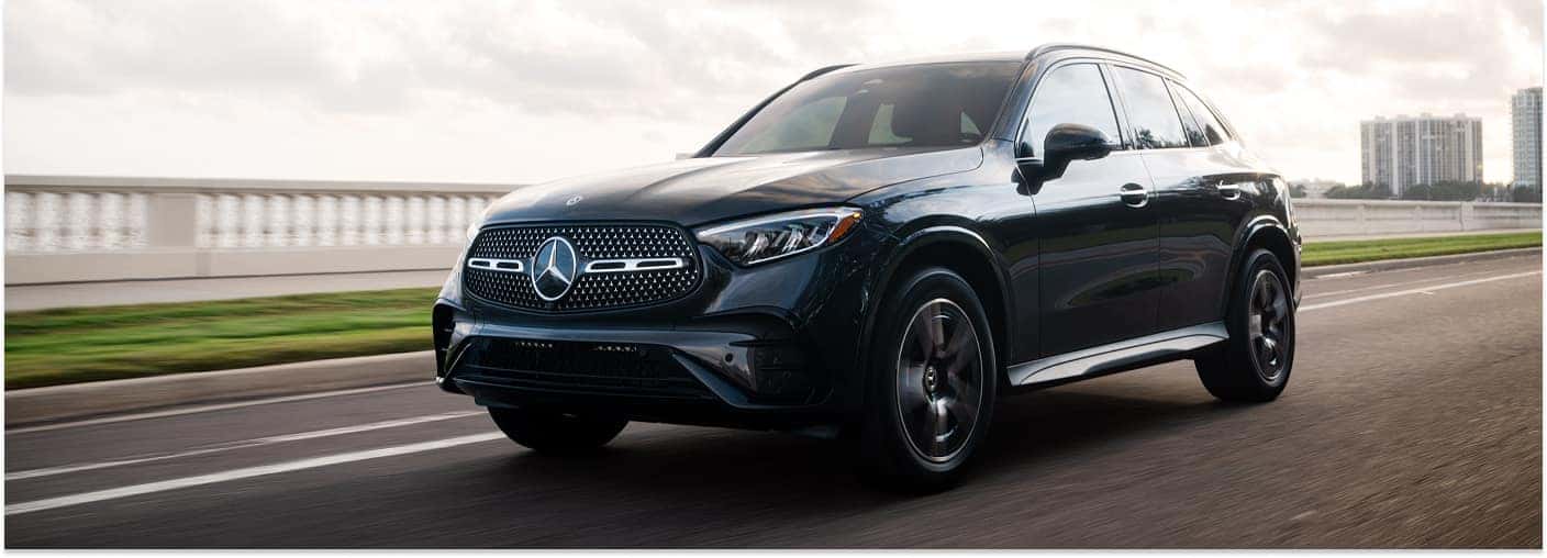 A front three-quarter view of a dark-colored 2026 Mercedes-Benz GLC SUV driving along a coastal road beside a white balustrade with city buildings in the background.