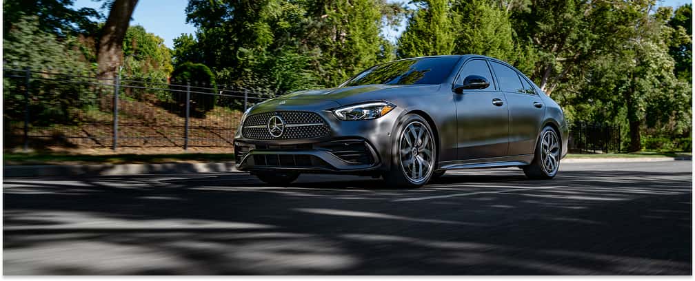 Front three-quarter view of a matte gray 2026 Mercedes-Benz C-Class sedan driving on a suburban street.