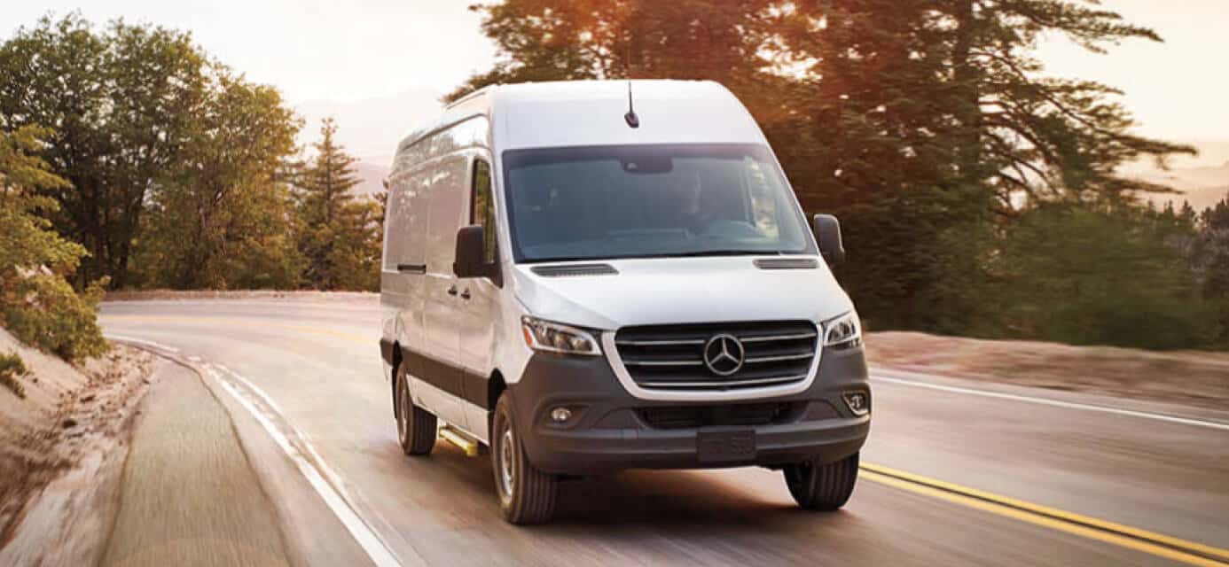 A white Mercedes Sprinter van cornering around a tree-lined road at golden hour.