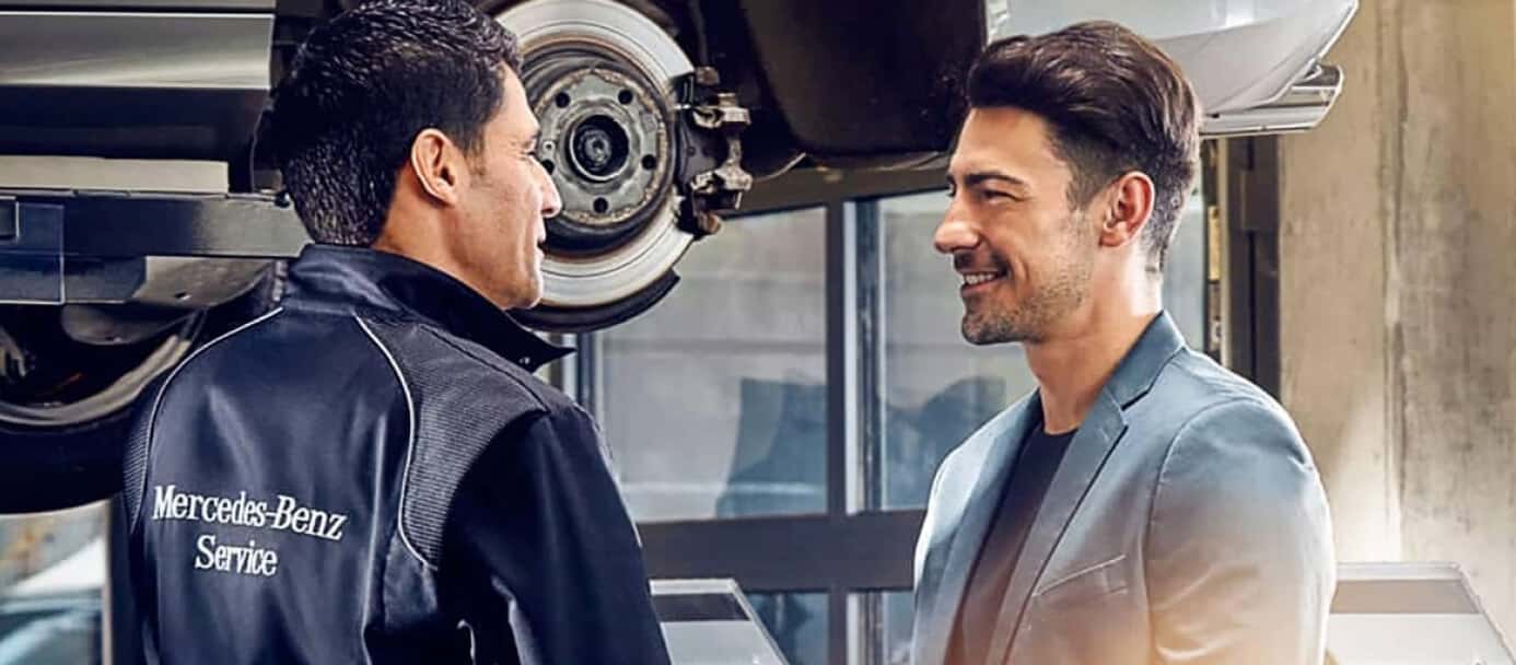 A Mercedes-Benz Service Technician speaks to a smiling customer near a lifted Mercedes car with the brake rotor exposed.