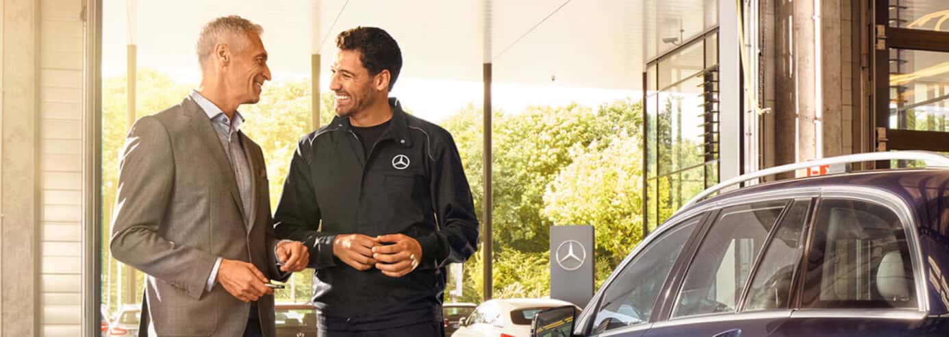 A Mercedes-Benz salesman stands with a smiling customer in a bright sunlit dealership near a new Mercedes vehicle.