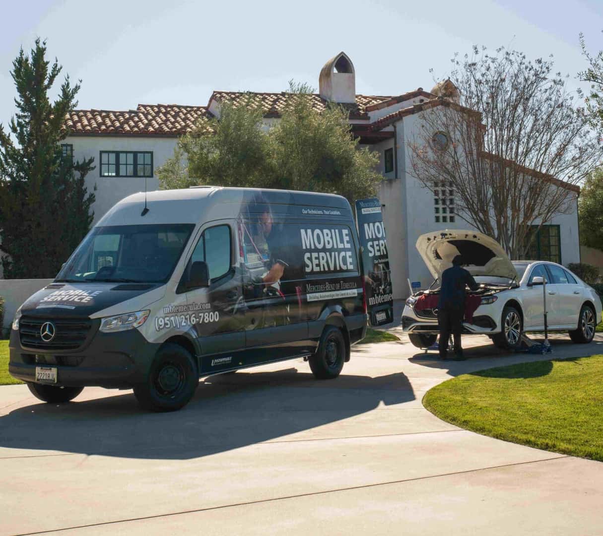 A Mercedes-Benz Mobile Service Van parked in the driveway of a home while a Service Technician inspects under the hood of a Mercedes car.