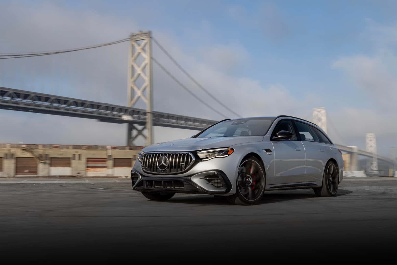 The High-Performing 2026 Mercedes AMG(R) E53 HYBRID Wagon Driving A Deserted Parking Lot, In View Of Storage Units And A Tall Bridge