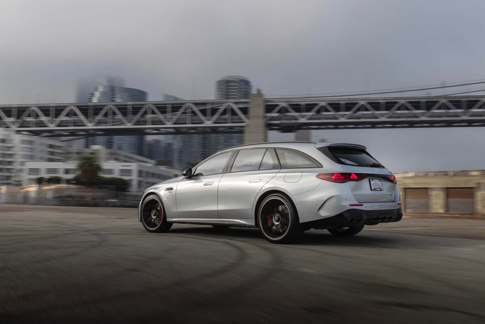 Rear Side Of The Stunningly Efficient The High-Performing 2026 Mercedes AMG(R) E53 HYBRID Wagon, Driving In View Of A Bridge And City Skyscrapers