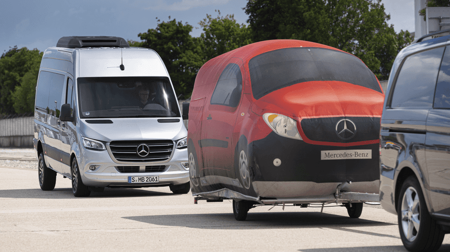 A Mercedes-Benz Sprinter Van behind a towing vehicle with a Mercedes-Benz art cover