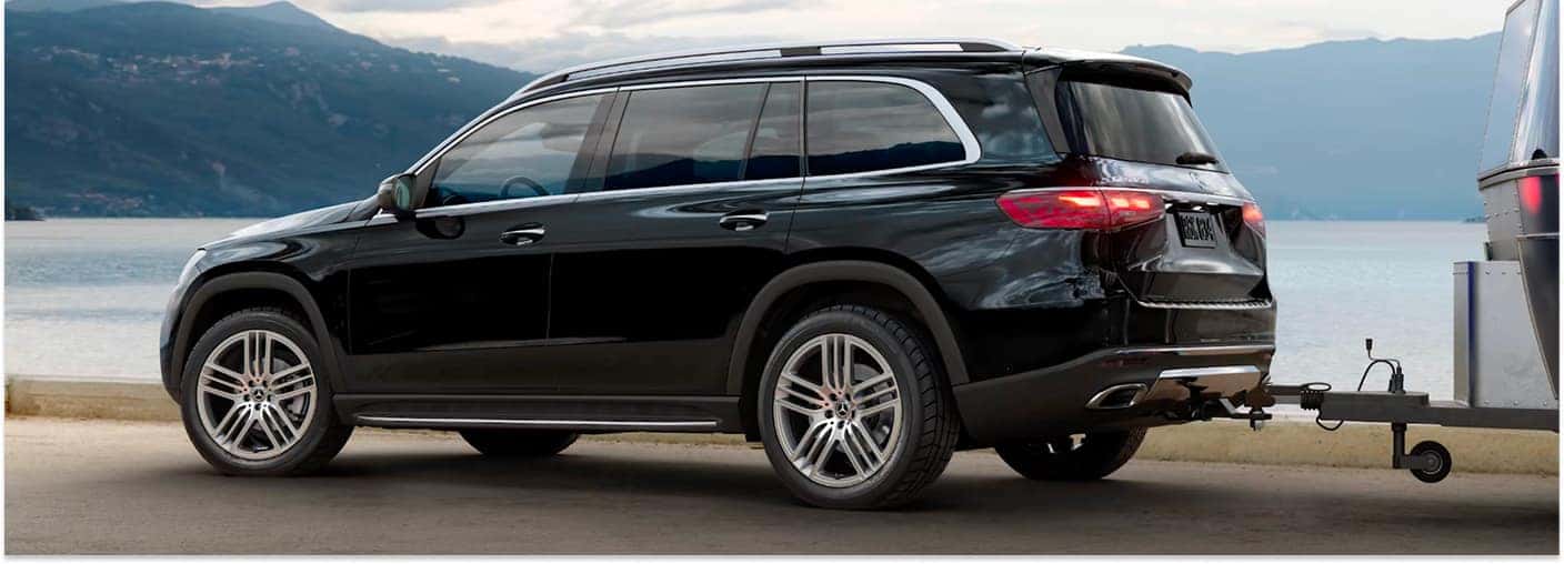 Black 2026 Mercedes-Benz GLS SUV towing a silver trailer parked by a lake, shown from a rear three-quarter view.