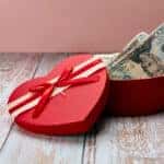 Gift in the shape of an open heart and inside full of dollars, on top of a wooden table and a pink background. Happy Valentine's Day, Mother's Day, birthday gift.