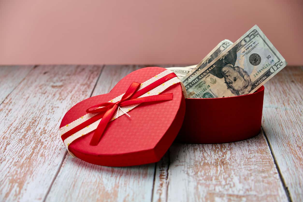 Gift in the shape of an open heart and inside full of dollars, on top of a wooden table and a pink background. Happy Valentine's Day, Mother's Day, birthday gift.