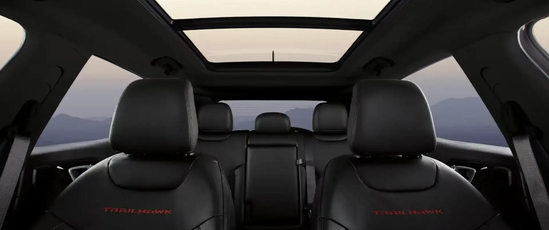 2026 Jeep Compass Trailhawk interior with black leather seats, red Trailhawk embroidery, and a panoramic sunroof framing a mountain sunset view