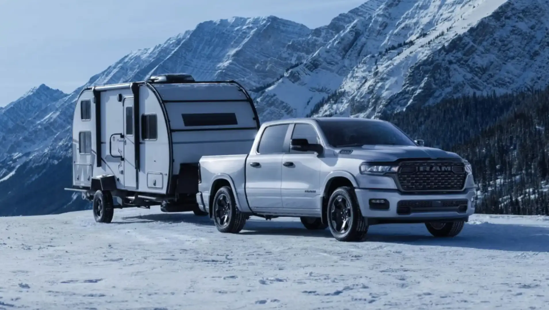 The 2026 Ram 1500 towing a small travel trailer across snowy terrain with snow-capped mountains in the background