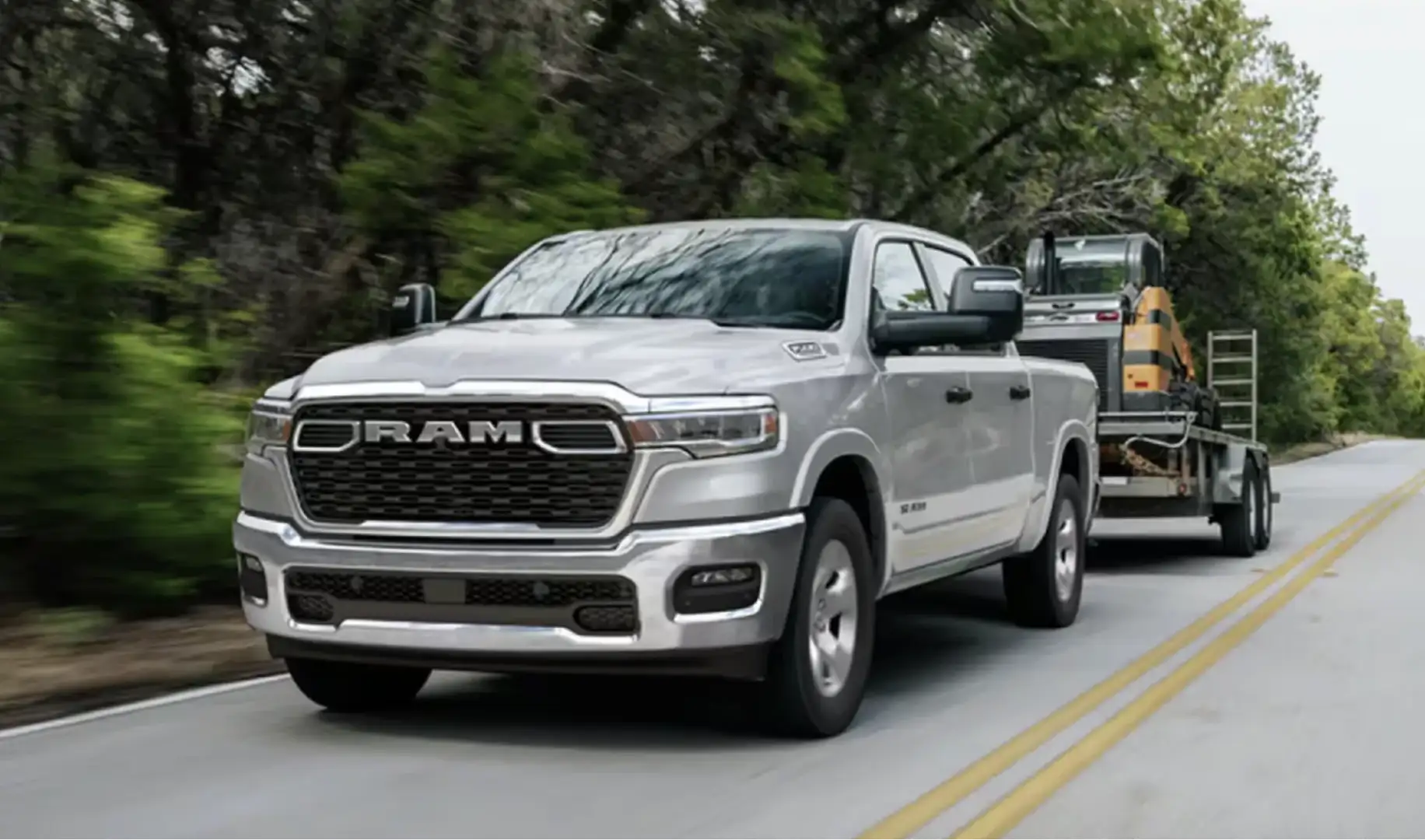 Ram 1500 pickup truck towing a flatbed trailer with construction equipment down a tree-lined road