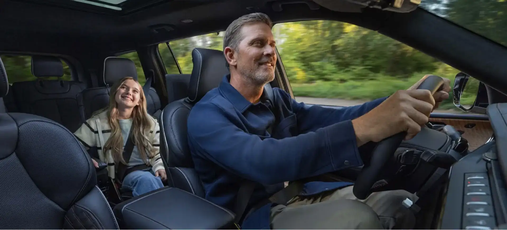 A smiling man drives a Jeep SUV down a tree-lined road while his young daughter rides happily in the back seat